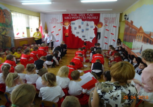 Na tle dekoracji z mapą Polski i dziećmi z chorągiewkami przedszkolaki kucają przygotowując się do tańca.
